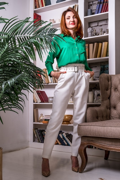 Una donna elegante in pantaloni bianchi e camicetta verde si erge sullo sfondo di una libreria in ufficio