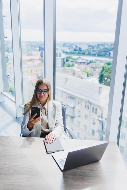 Una donna europea allegra dai capelli biondi con gli occhiali in abiti casual eleganti è seduta a un tavolo con un laptop che fa scartoffie e parla al telefono Signora d'affari sul posto di lavoro in ufficio