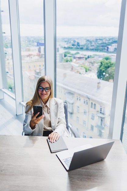 Una donna europea allegra dai capelli biondi con gli occhiali in abiti casual eleganti è seduta a un tavolo con un laptop che fa scartoffie e parla al telefono Signora d'affari sul posto di lavoro in ufficio