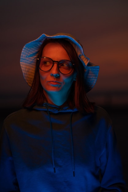 Una donna europea illuminata da una luce al neon con un cappello sullo sfondo del cielo al tramonto