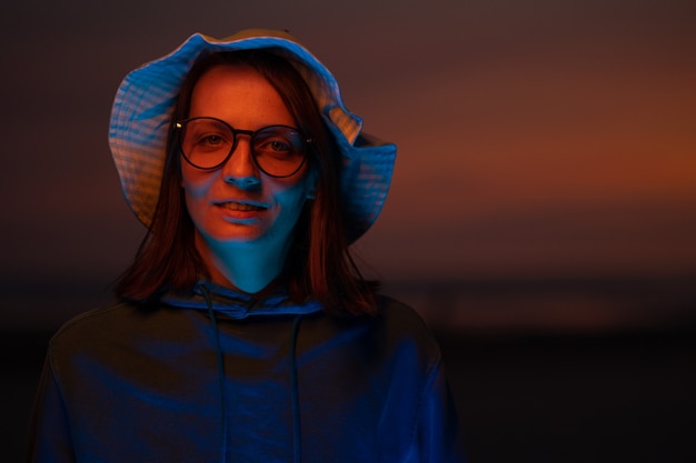 Una donna europea illuminata da una luce al neon con un cappello sullo sfondo del cielo al tramonto