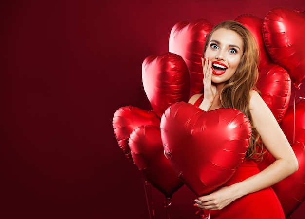 Una donna felice con un vestito rosso con dei palloncini un cuore rosso su uno sfondo di bandiera rossa una ragazza allegra e sorpresa con il trucco sulle labbra rosse e un sorriso carino una sorpresa per la gente di San Valentino e il concetto del giorno di Sant'Valentino