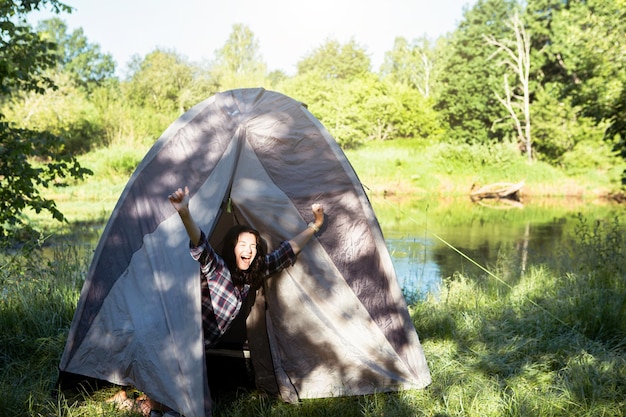 Una donna felice con una camicia a quadri guarda fuori da una tenda turistica durante un'escursione sulla riva del fiume al mattino Campeggio nella natura pernottamento nella natura selvaggia vacanze e avventure in famiglia