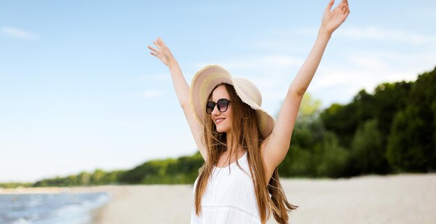 Una donna felice e sorridente in felicità libera sulla spiaggia dell'oceano in piedi con un cappello, occhiali da sole e mani rabbiose. Ritratto di una modella femminile multiculturale in abito bianco d'estate che si gode la natura