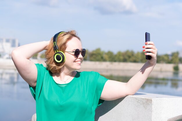 Una donna felice in occhiali da sole e cuffie che scatta una foto selfie sul telefono che chatta con gli amici per strada