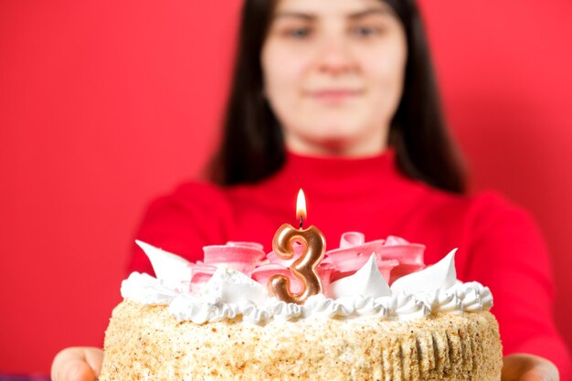 Una donna felice tiene una torta con una candela a forma di numero tre su sfondo rosso