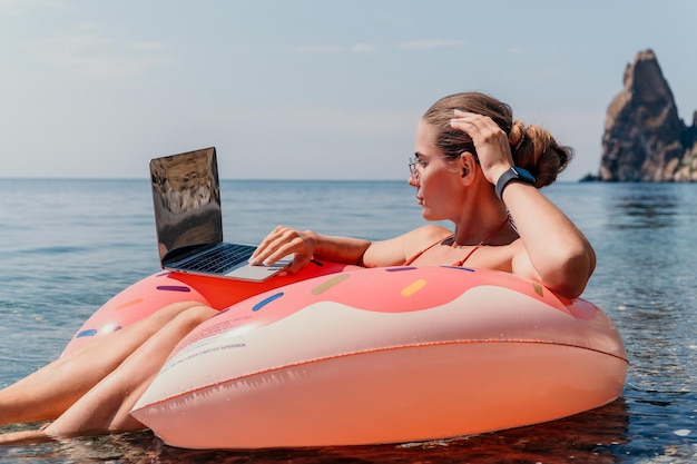 Una donna freelance lavora su un computer portatile nuotando in mare su un anello gonfiabile rosa, un turista felice che galleggia sopra