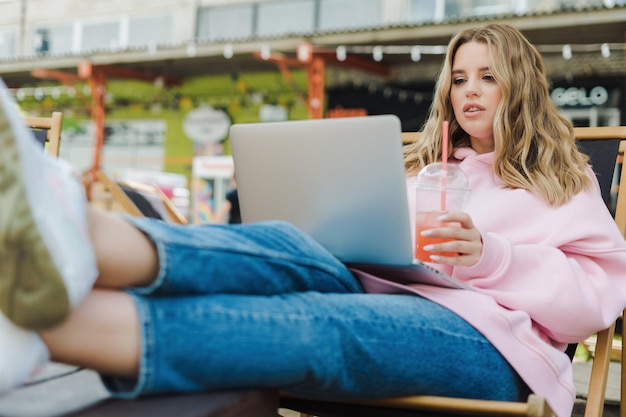 Una donna freelance lavora su un laptop e beve limonata in un caffè di strada Una modella con una felpa con cappuccio rosa e jeans