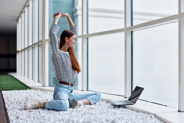 Una donna freelance si siede nella hall dell'ufficio vicino alle grandi finestre con un portatile online tramite Internet freelance lavoro ovunque nel mondo
