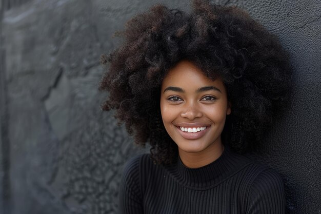 Una donna gioiosa con un afro voluminoso sorride brillantemente guardando la telecamera appoggiata a un muro nero della città concetto di atteggiamento positivo