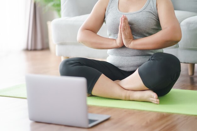 Una donna grassa e paffuta asiatica matura seduta sul pavimento in soggiorno pratica lezioni di yoga online con il computer. femmina che ha meditare la lezione di formazione sul laptop.