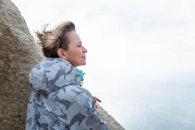 Una donna guarda il mare mentre sta in piedi su una montagna in una giornata di sole Una carina bionda con una giacca grigia Ricreazione attiva in natura Closeup