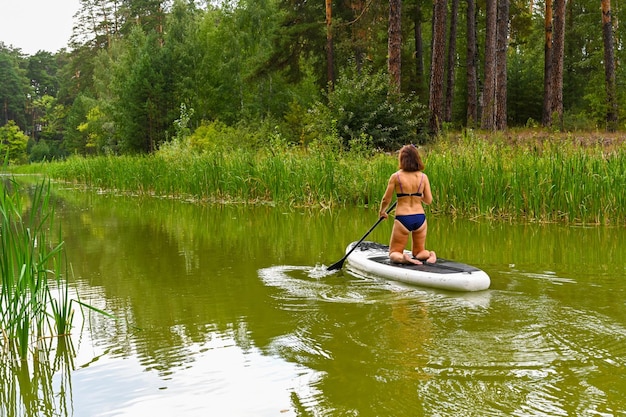 Una donna guida sulla Sup Board attraverso uno stretto canale circondato da erba fitta Vacanze attive per il fine settimana natura selvaggia all'aperto La donna è in piedi in grembo in costume da bagno