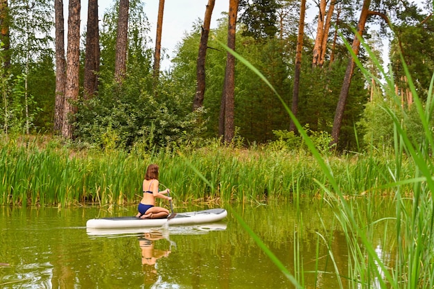 Una donna guida sulla tavola da Sup attraverso uno stretto canale circondato da erba fitta. Vacanze attive per il fine settimana natura selvaggia all'aperto. La donna è seduta sulle ginocchia in costume da bagno. L'estate.