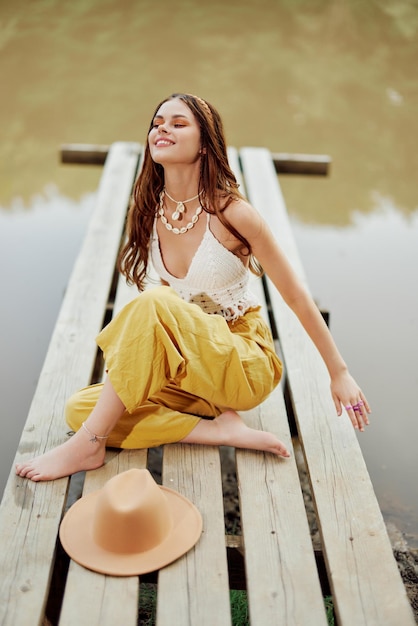 Una donna hippie siede con il suo cappello su un ponte vicino a un lago con le mani alzate durante un viaggio nella natura e sorride felicemente in ecoabbigliamento Stile di vita rilassato