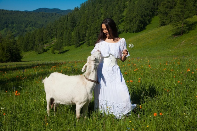 Una donna in abito bianco con una capra bianca cammina sui monti Altai in estate.