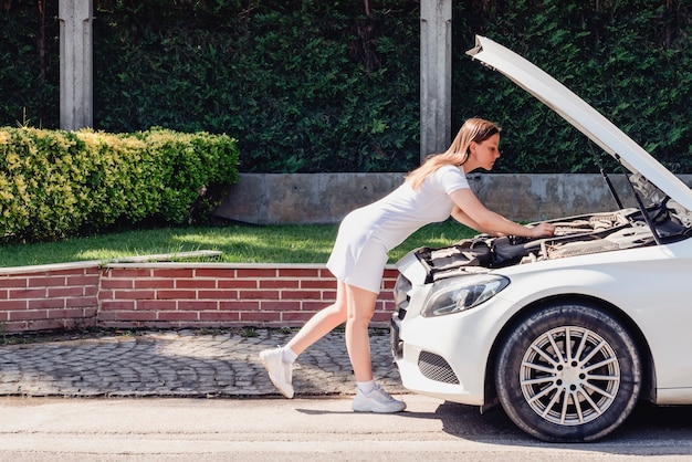 Una donna in abito bianco sta cercando di riparare da sola la sua auto bianca che si è rotta sul fi...