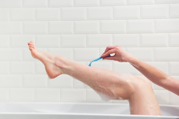 Una donna in bagno si rade le gambe con un rasoio. Primo piano di una mano con un rasoio.