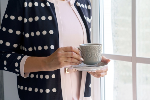 Una donna in cardigan scuro è in piedi vicino alla finestra con un berretto in mano