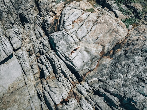 Una donna in costume da bagno nero sdraiata su una calda costa rocciosa fratturata, ripresa da un drone, sfondo.