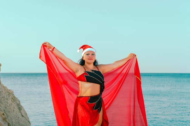 Una donna in costume per danze orientali con un cappello da Babbo Natale sullo sfondo del mare e delle rocce sta ballando con un velo. Foto orizzontale