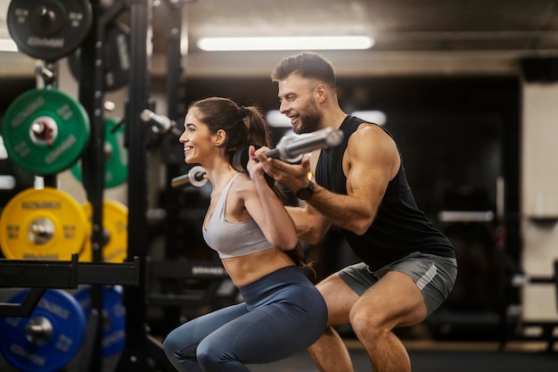 Una donna in forma si sta accovacciando con un bar in una palestra mentre il suo allenatore la aiuta