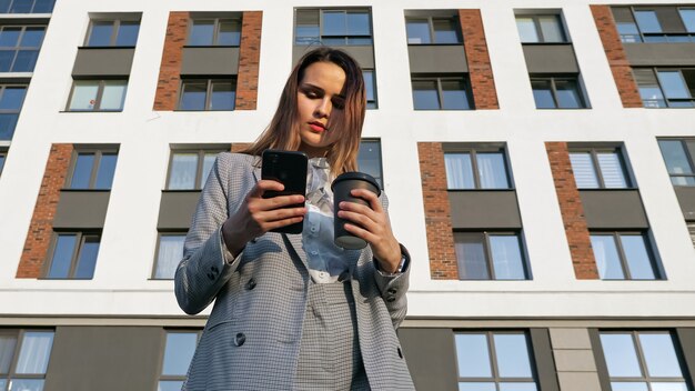 Una donna in giacca e cravatta beve caffè da un bicchiere di plastica e guarda il telefono sullo sfondo di un edificio