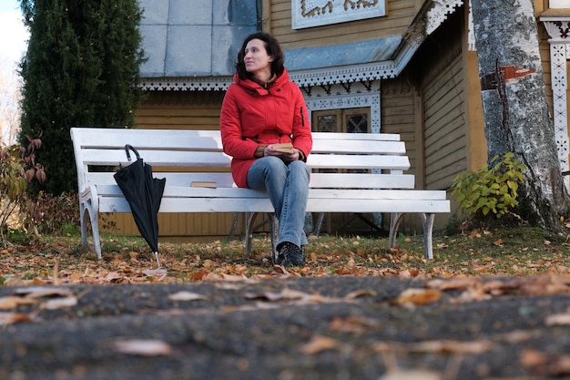 Una donna in giacca rossa è seduta su una panchina bianca nel parco e legge un libro di carta.