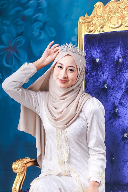 Una donna in hijab con una corona in testa si trova di fronte a un muro blu.