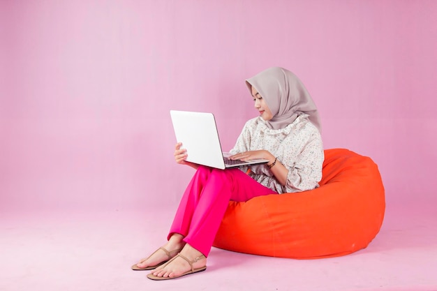 Una donna in hijab siede su un pouf e lavora su un computer portatile.