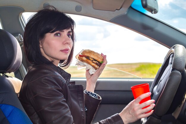 Una donna in macchina con un hamburger e un bicchiere in mano