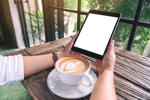 una donna in possesso di tablet pc nero con schermo desktop bianco bianco con una tazza di caffè sul tavolo di legno