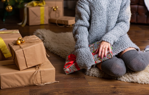 Una donna in un abito grigio lavorato a maglia si siede sul tappeto e tiene in mano un regalo alla vigilia di Natale