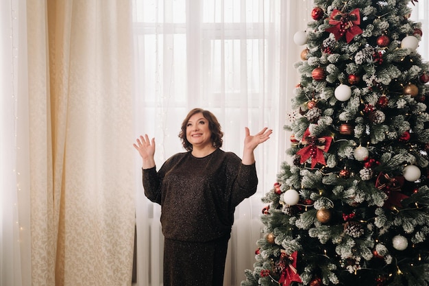 Una donna in un abito scuro in piedi accanto a un albero di Natale in un interno progettato per celebrare il Natale e il Capodanno