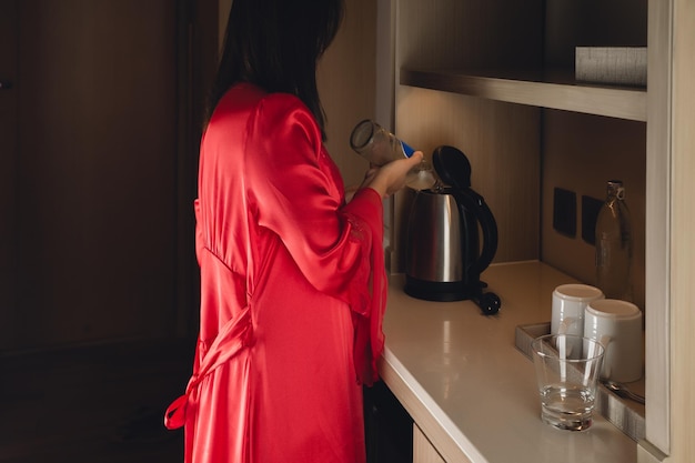 Una donna in una camicia da notte di raso versa acqua pulita da una bottiglia in un bollitore in cucina di notte