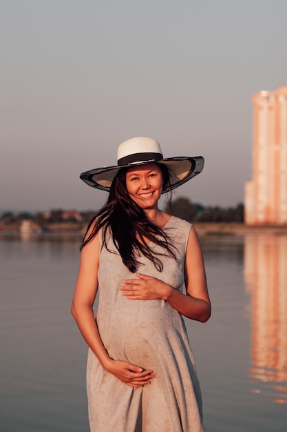 Una donna incinta al tramonto una donna incinta sorridente felice con un cappello di paglia e un vestito grigio la tiene ...