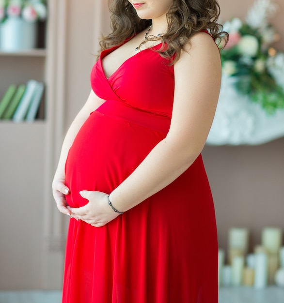 Una donna incinta con i capelli scuri in un vestito rosso abbraccia teneramente la sua grande pancia con le braccia. Ritaglia