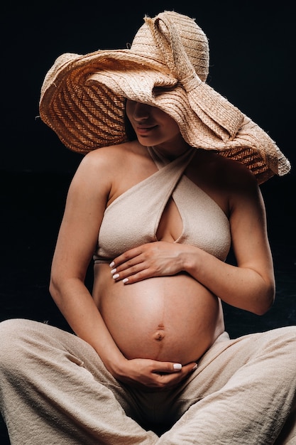 Una donna incinta con un cappello di paglia si siede sul pavimento in abiti beige in uno studio su sfondo nero.