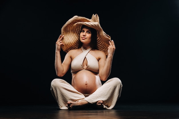 Una donna incinta con un cappello di paglia si siede sul pavimento in abiti beige in uno studio su sfondo nero.