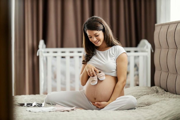 Una donna incinta è seduta su un letto con le scarpe da bambino in mano e la pancia coccolata