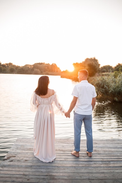 Una donna incinta e suo marito stanno sul molo e si guardano un bellissimo fiume e al tramonto i futuri genitori trascorrono del tempo all'aperto una bellissima giovane madre