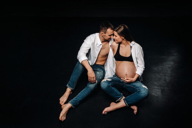 Una donna incinta e un uomo in camicia bianca e jeans in uno studio su sfondo nero.