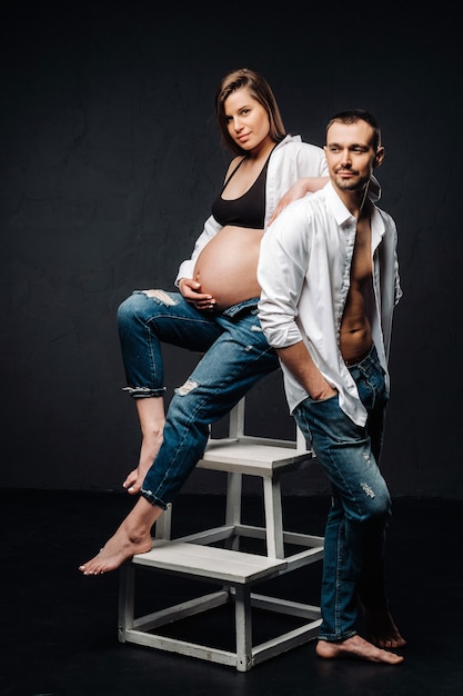 Una donna incinta e un uomo in camicia bianca e jeans in uno studio su sfondo nero.