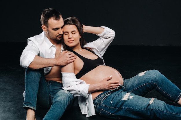 Una donna incinta e un uomo in camicia bianca e jeans in uno studio su sfondo nero.