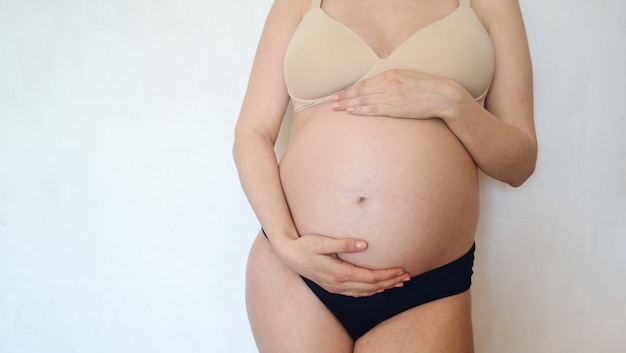 Una donna incinta in lingerie si erge sullo sfondo di un muro bianco e si accarezza la pancia. Concetto - maternità e gravidanza.