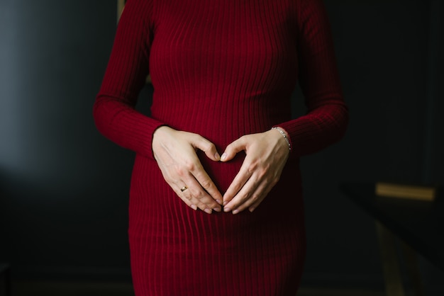 Una donna incinta in un abito lavorato a maglia bordeaux tiene le mani sul suo stomaco a forma di cuore