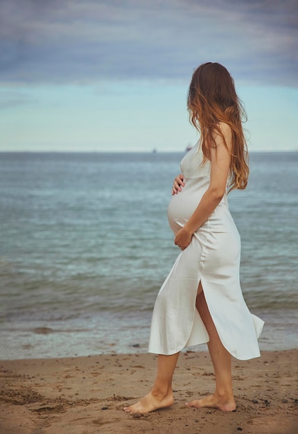 Una donna incinta in un vestito guarda il mare. Foto verticale