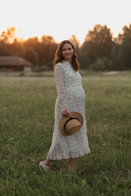 Una donna incinta su uno sfondo al tramonto è in posa e tiene il cappello con la mano vicino alla fattoria