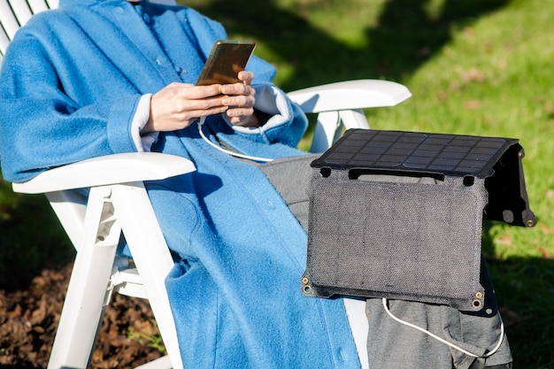 Una donna irriconoscibile con un telefono cellulare in mano e una batteria solare portatile siede all'aperto nel giardino Concetto ecologico di energia alternativa