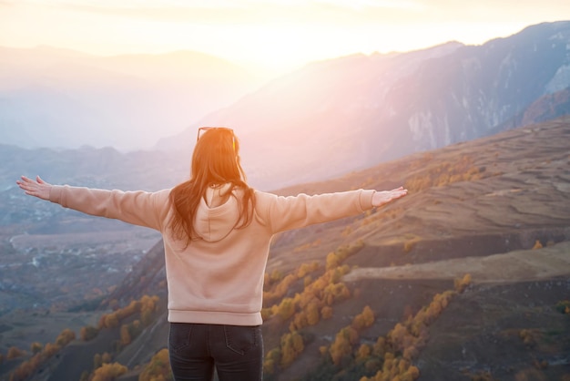 Una donna irriconoscibile con una felpa con cappuccio sta con le braccia aperte su uno sfondo di montagne, luce del sole.